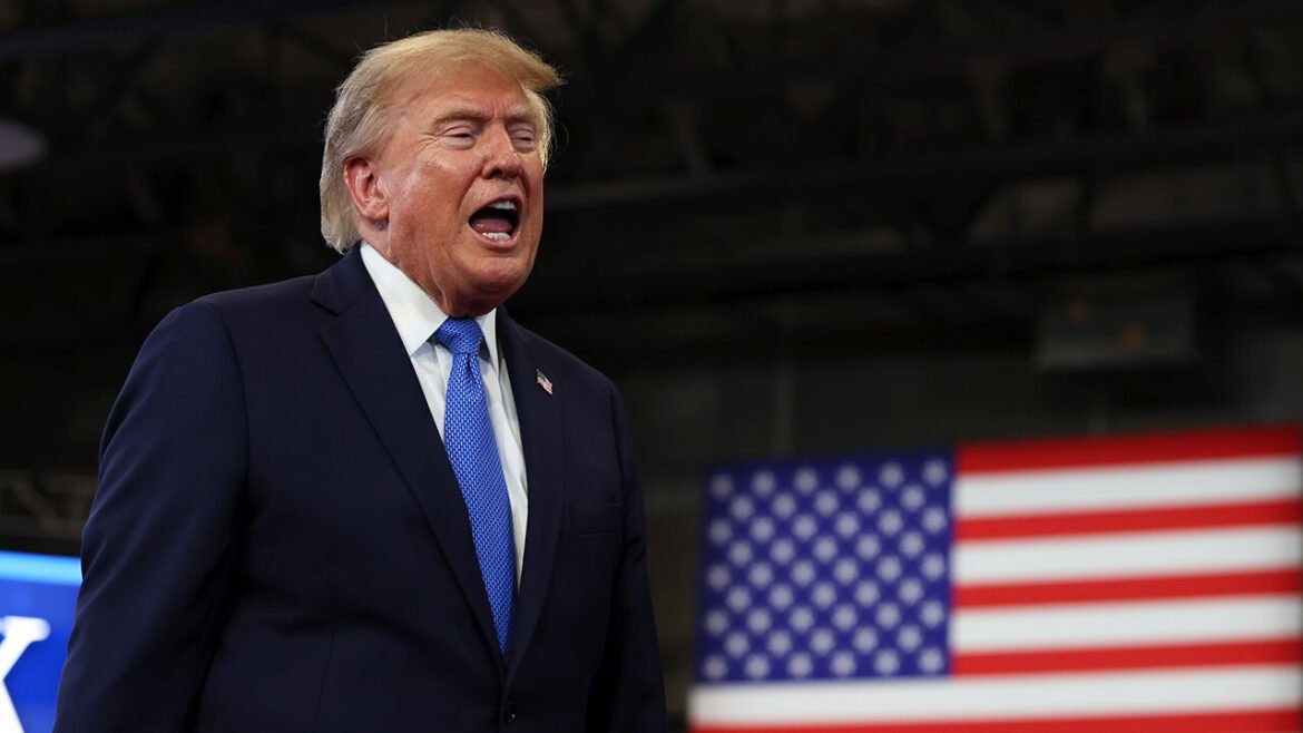 Man in a dark suit and blue tie speaking, with a large American flag in the background.