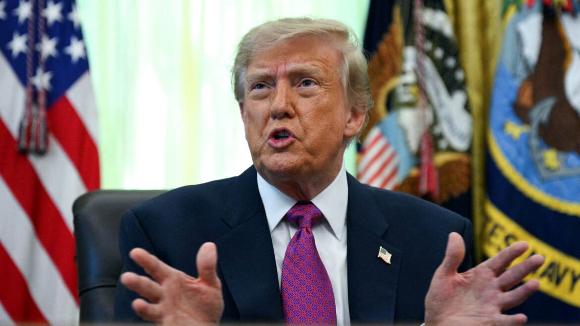 Man in a dark suit and red tie speaks, hands raised, with American flags in the background nearby flags behind him.