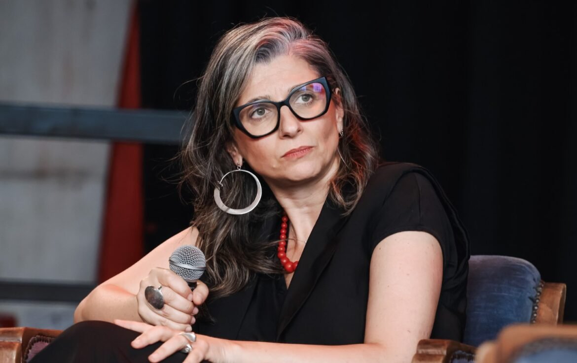 Woman with dark hair and glasses holds a microphone, listening thoughtfully during a panel discussion.