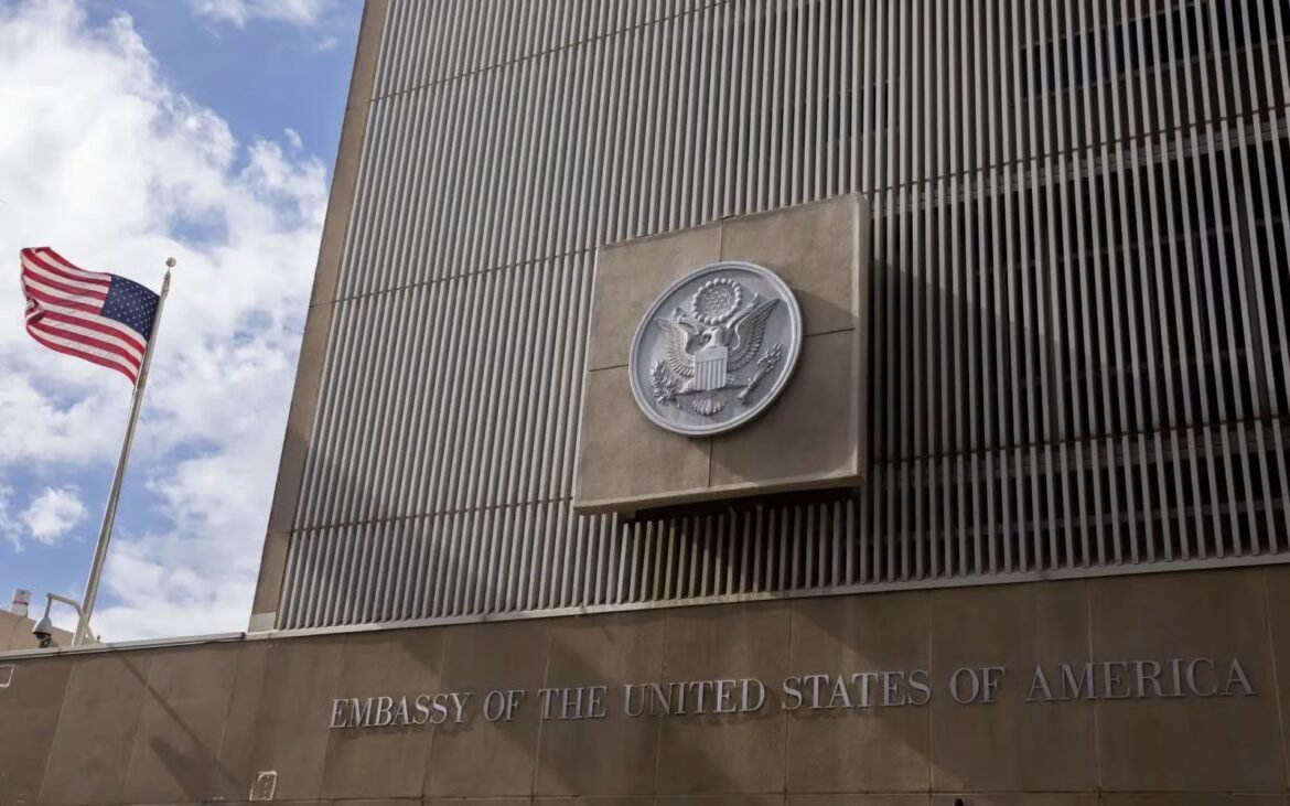 U.S. flag hoisted beside a modern embassy facade with a large circular seal mounted on the wall and the text 'EMBASSY OF THE UNITED STATES OF AMERICA' below.