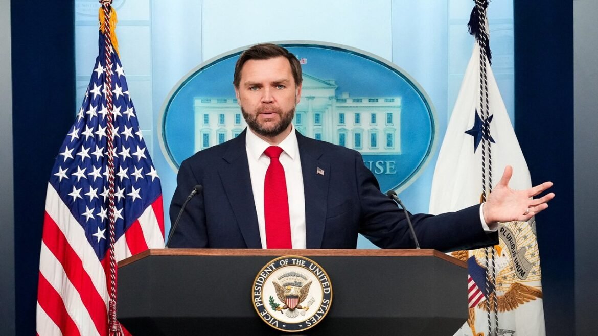 Man in a dark suit and red tie speaks at a podium with the presidential seal; American flag on left and a government flag on the right in a briefing room backdrop.