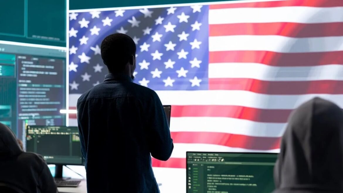 Person standing in a tech workspace, facing a wall-sized American flag displayed beside multiple monitors and code on screens.