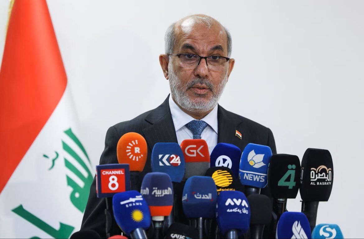 Man in a suit and glasses speaking at a press conference with multiple microphones in front of him.
