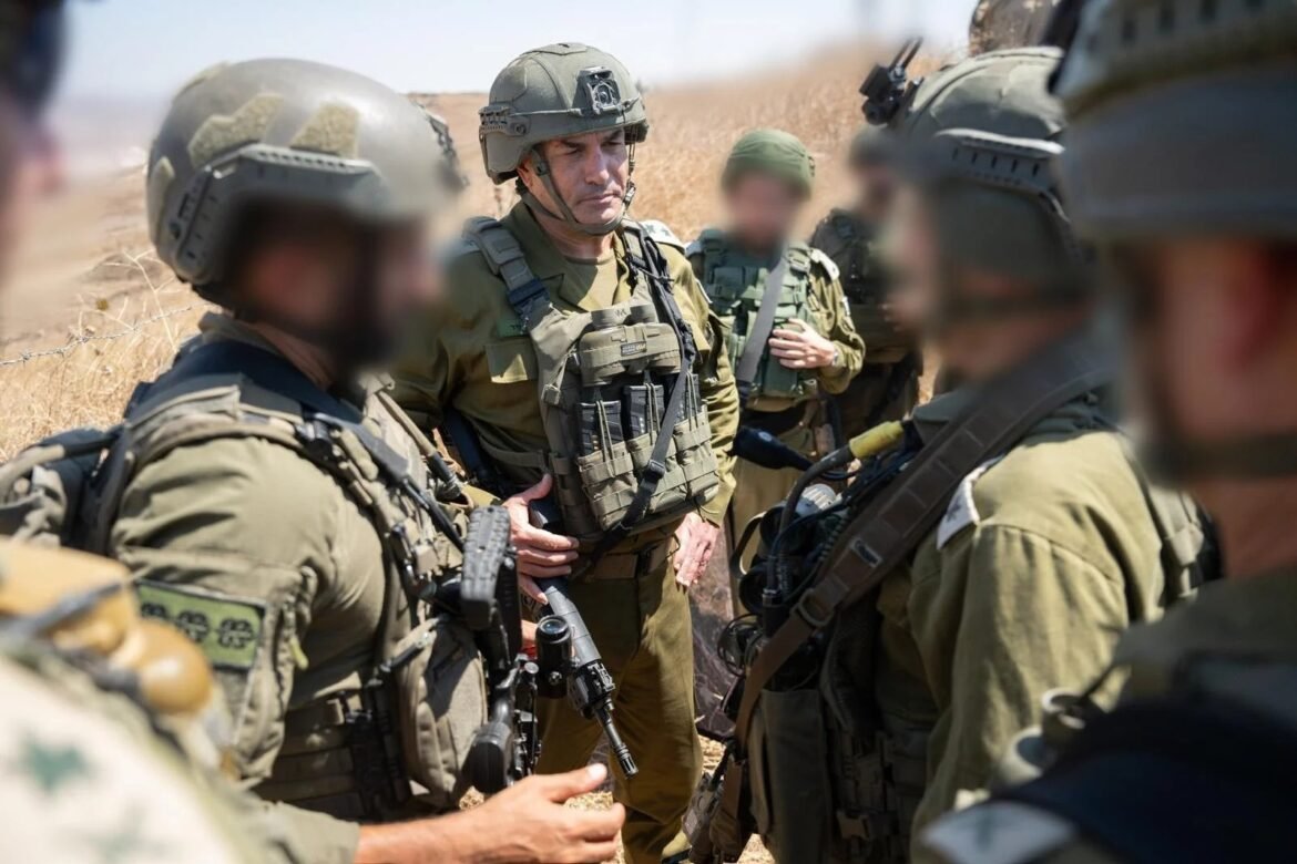 Soldiers in full combat gear standing in a circle, listening during a field briefing in a dry, grassy area.