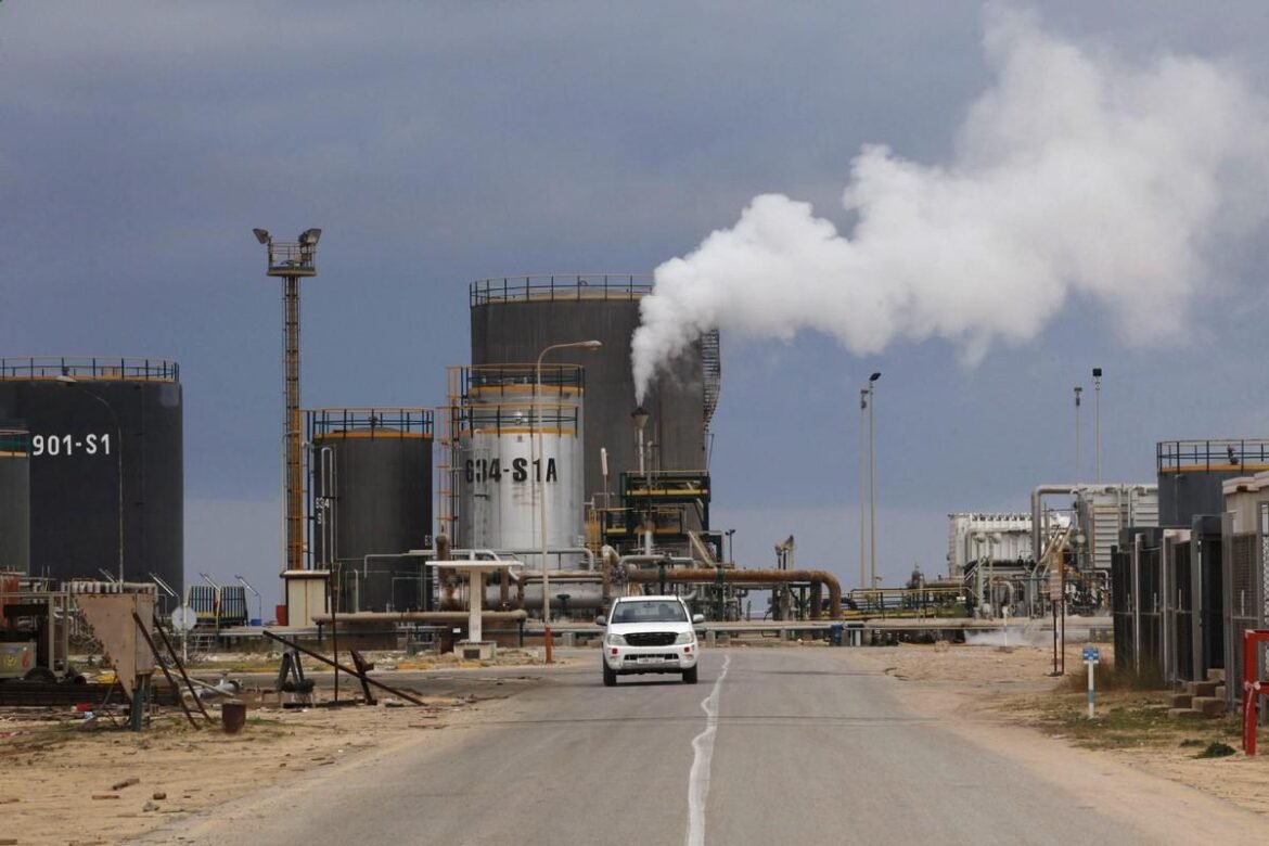 White car drives toward an industrial refinery with large storage tanks and steaming chimneys on a dusty road under a cloudy sky