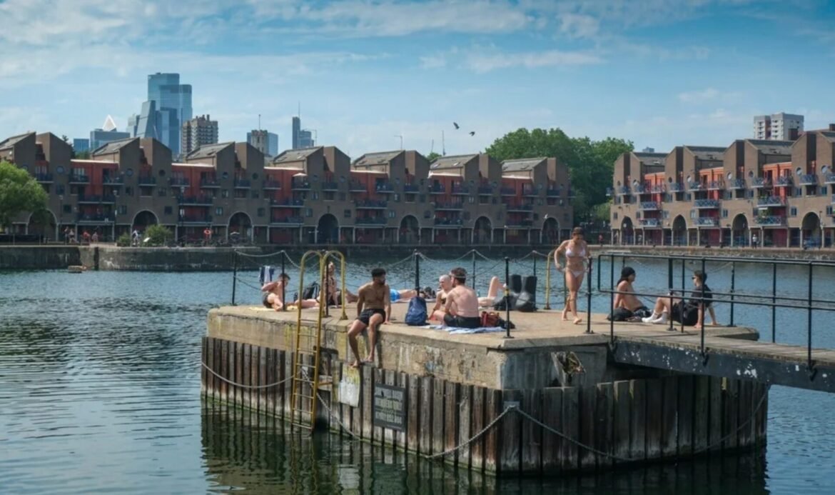 People sunbathing on a concrete pier by a calm harbor with a city skyline in the background, blue sky above.