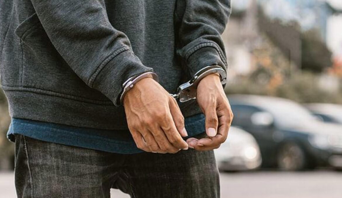 Person in a dark gray hoodie with metal handcuffs on their wrists, standing outdoors.