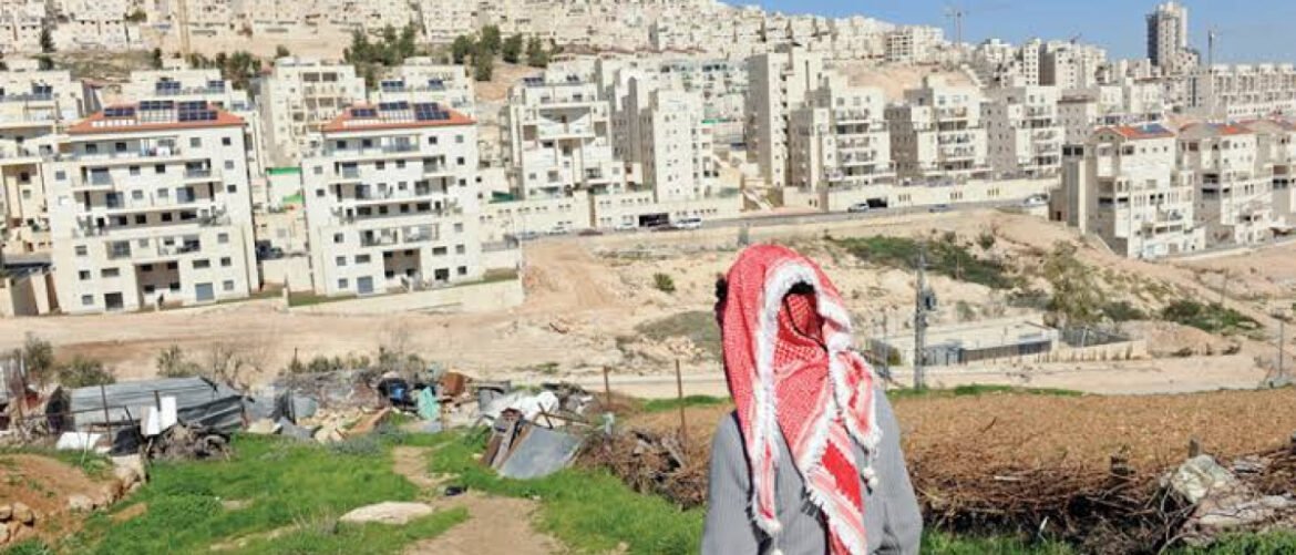 Person wearing a red-and-white keffiyeh facing a dense city of beige apartment blocks, standing on rubble-strewn ground in the foreground.