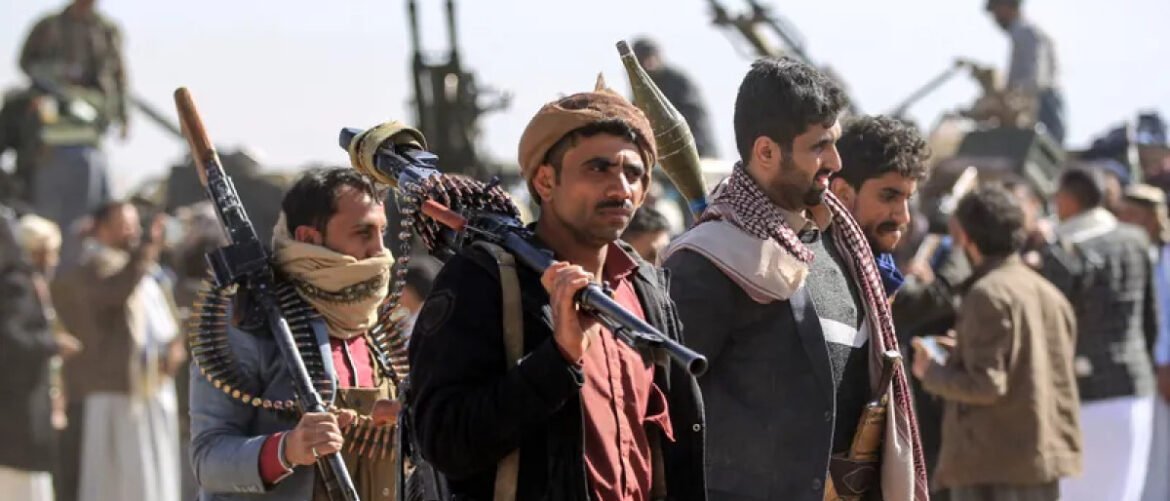 Group of men with rifles and ammo belts standing in a crowd, some wearing scarves and traditional clothing in a dusty outdoor setting.