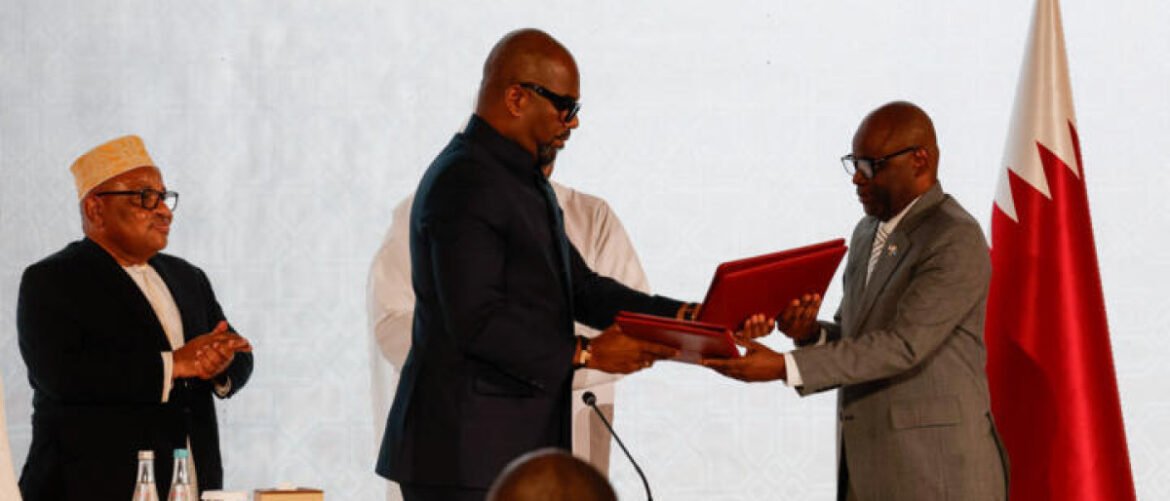Two men in formal suits exchange red folders at a ceremonial event, while another man applauds and a Qatari flag stands nearby.