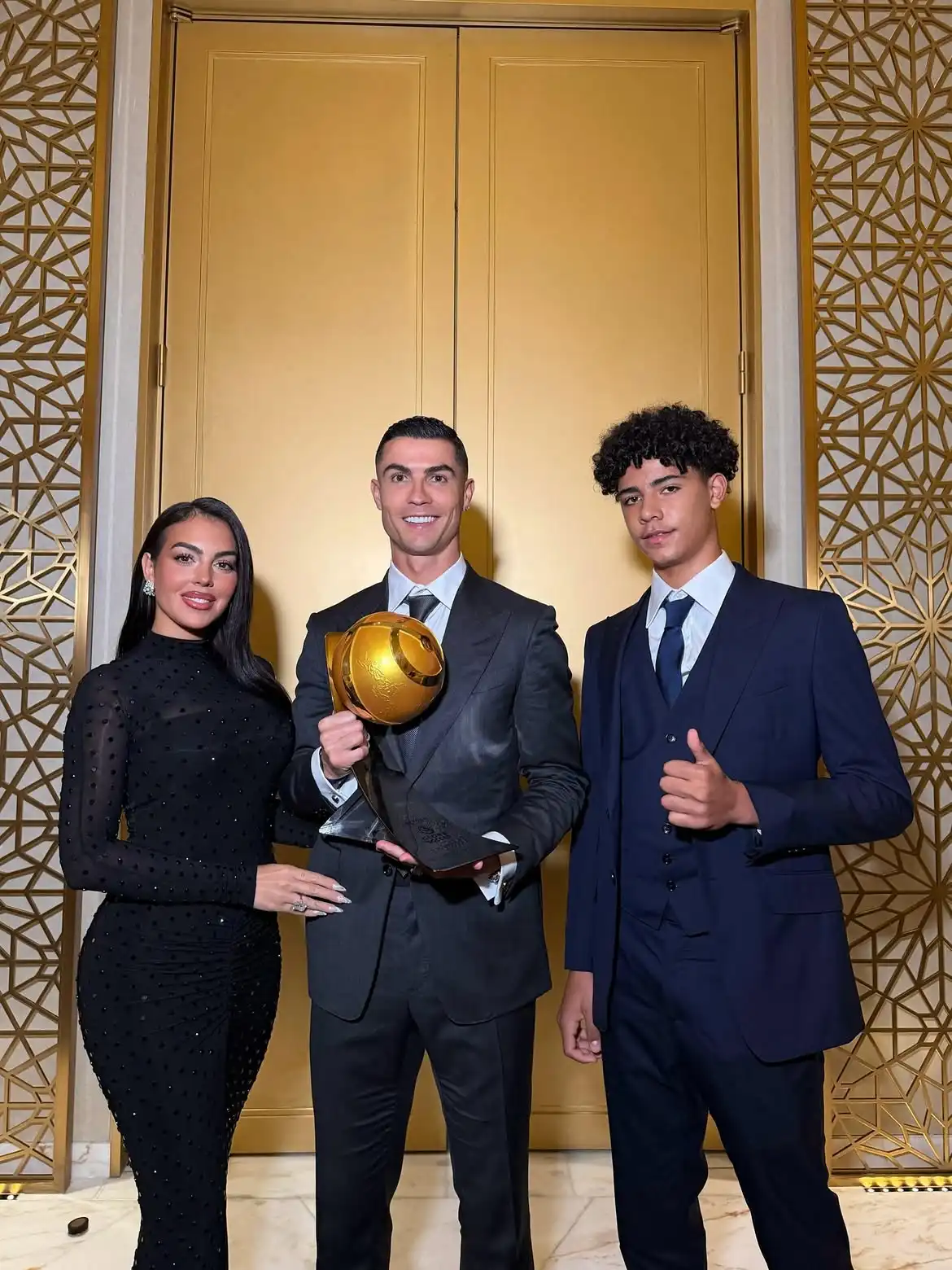 Three People in Formal Attire Pose with a Golden Ballon Dor Trophy in Front of Ornate Gold Doors in a Stylish Venue  Aleumumnews