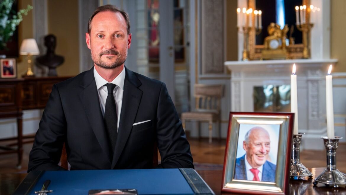 Man in a dark suit sits at a desk in a formal room with framed photos and lit candles in the background.