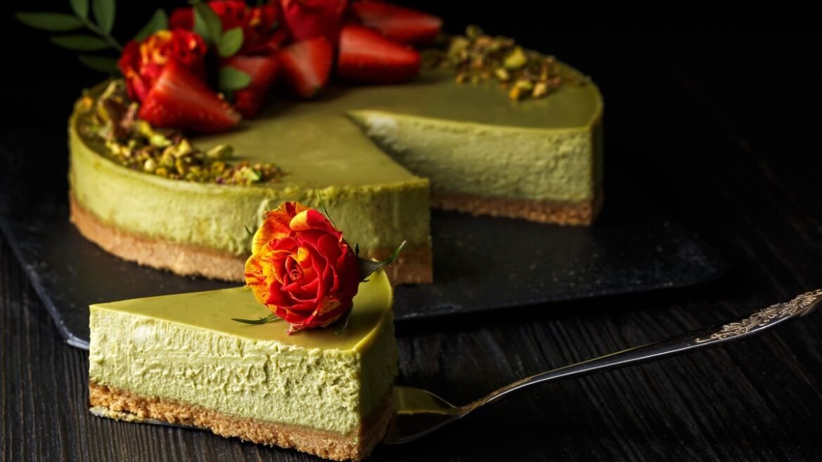 Slice of pale green pistachio cheesecake with a crumb crust and a red-orange rose on top, on a dark wooden table.