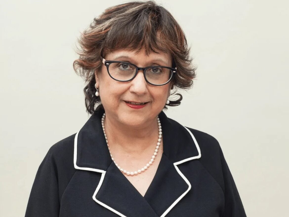 Middle-aged woman in a black blazer with white piping, pearl necklace, and glasses, smiling at the camera in a professional head-and-shoulders portrait.