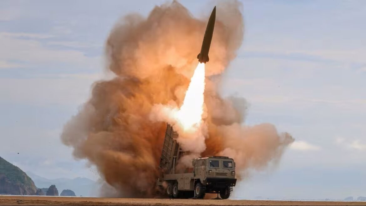 Missile launch from a military transporter on a sandy beach with a bright flame and orange smoke plume behind it.