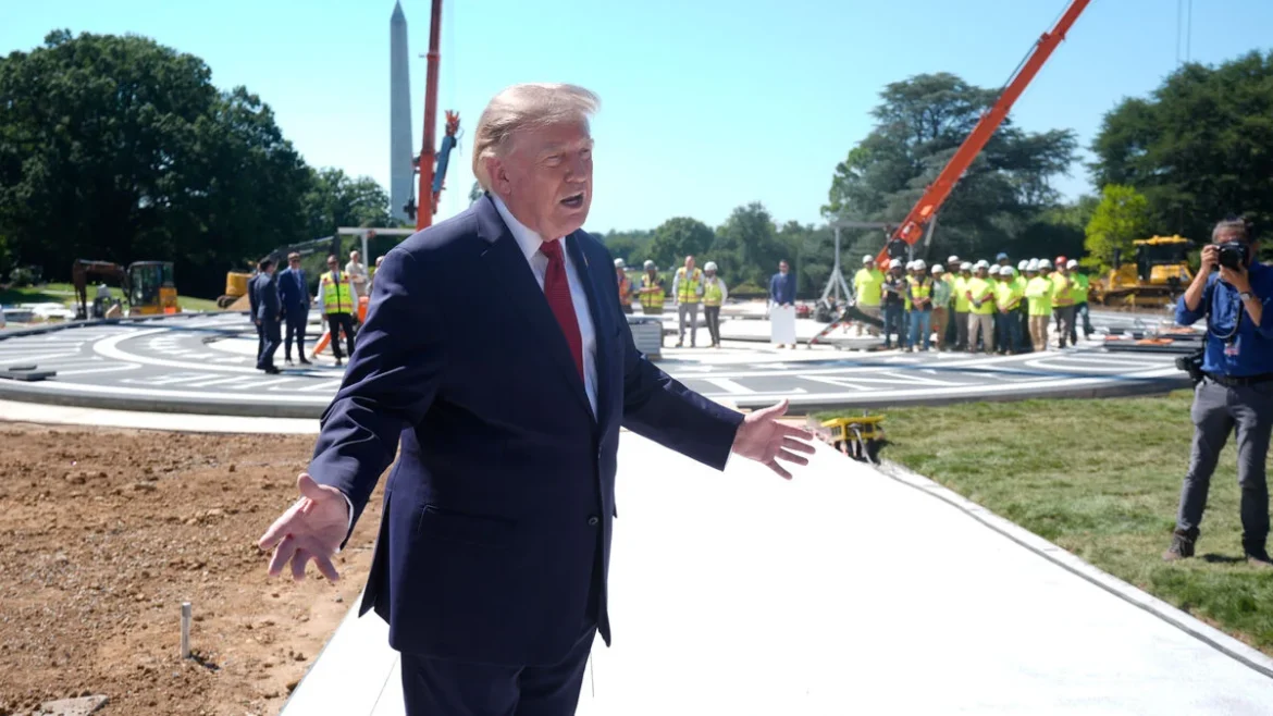 Man in a dark suit speaks with arms open at a construction site, with workers, cranes, and a large circular platform behind him on a sunny day.