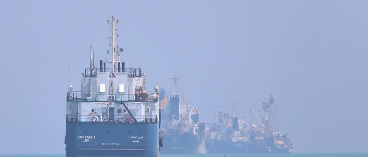 Convoy of blue cargo ships anchored in a hazy harbor, with the closest vessel in the foreground.