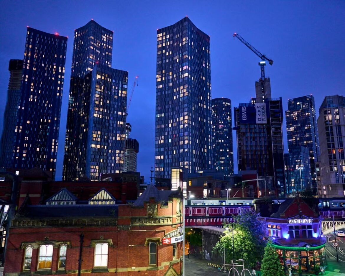 Night city skyline at blue hour with tall blue glass towers, a construction crane, and a red-brick station in the foreground.