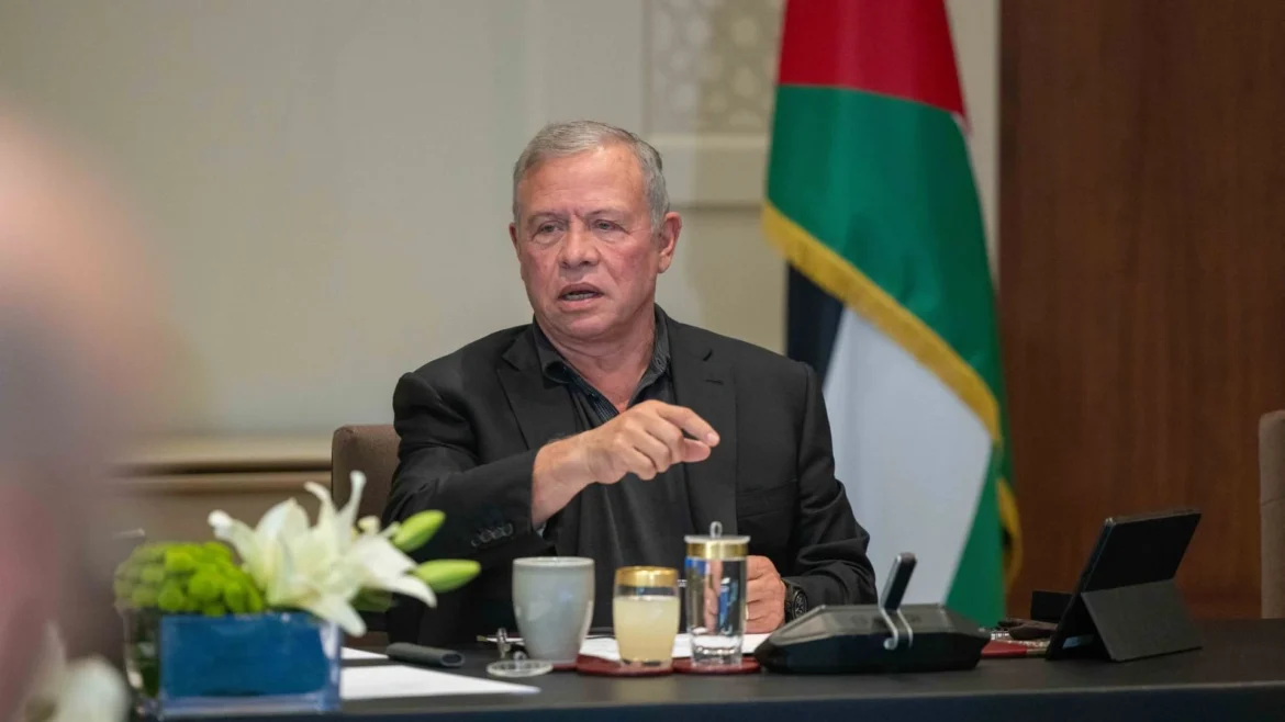 Man in a dark suit speaks at a conference table during a formal meeting, with a flag in the background and drinks on the table behind him.