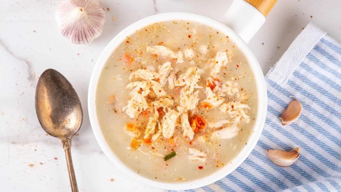 Creamy chicken soup in a white bowl topped with shredded chicken and spices, with garlic cloves, a spoon, and a blue striped cloth nearby.