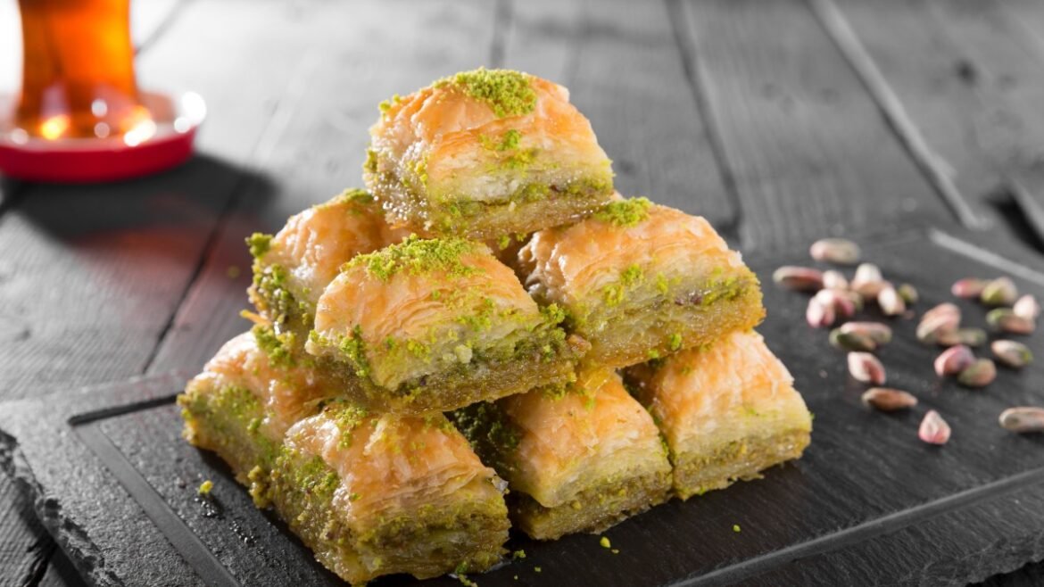 Stack of pistachio baklava squares stacked on a dark slate with chopped pistachios and a glass of tea in the background