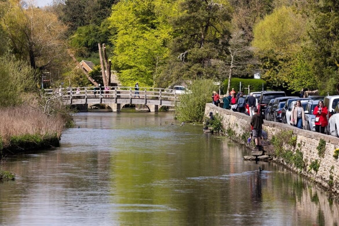 People walk along a riverside path beside a stone wall and parked cars, with a wooden bridge over calm water in a park setting.