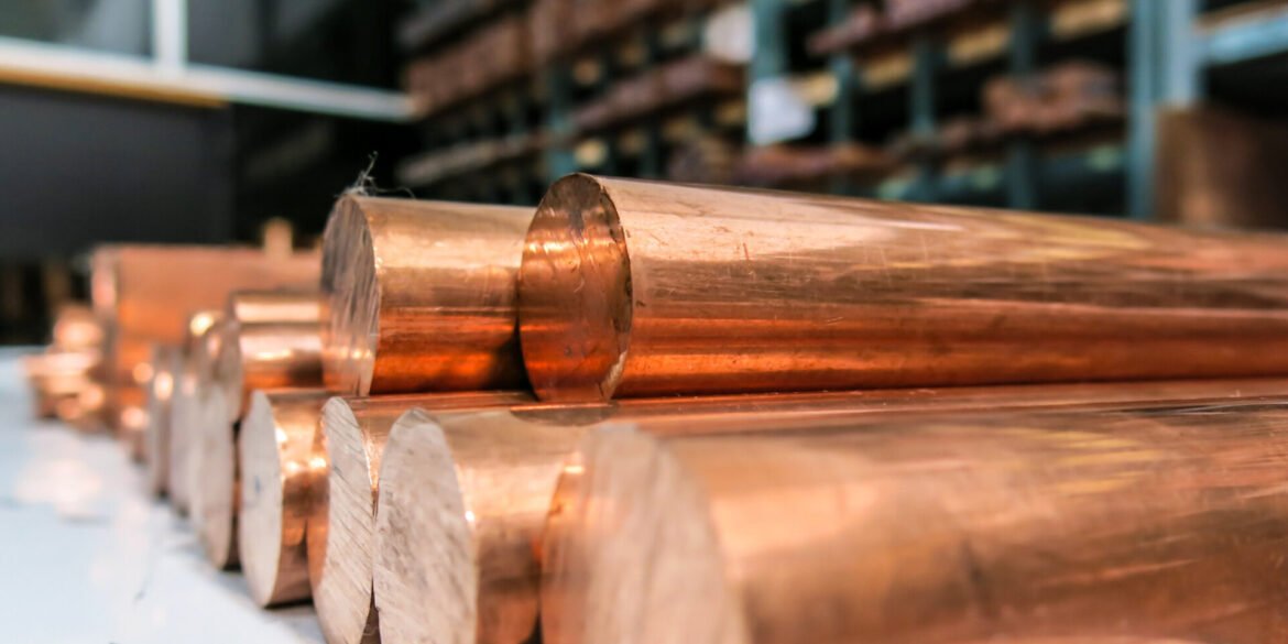 Copper cylindrical metal rods stacked in a warehouse, with warm reflective surfaces and a blurred background.