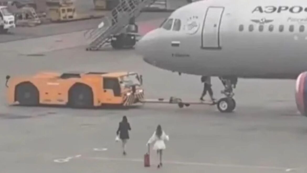 Airplane on the tarmac being pushed back by an orange tug; boarding stairs nearby and a few passengers walking in the foreground.