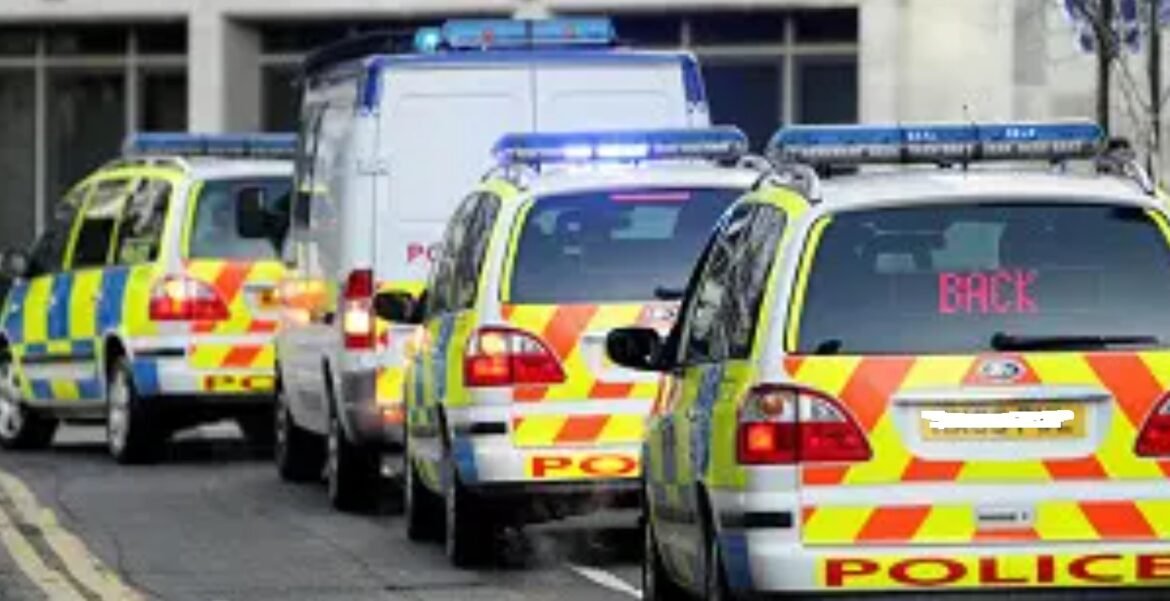 Line of police cars with bright yellow/red chevrons and blue lights, forming a convoy on a city street, one labeled BACK on rear window.
