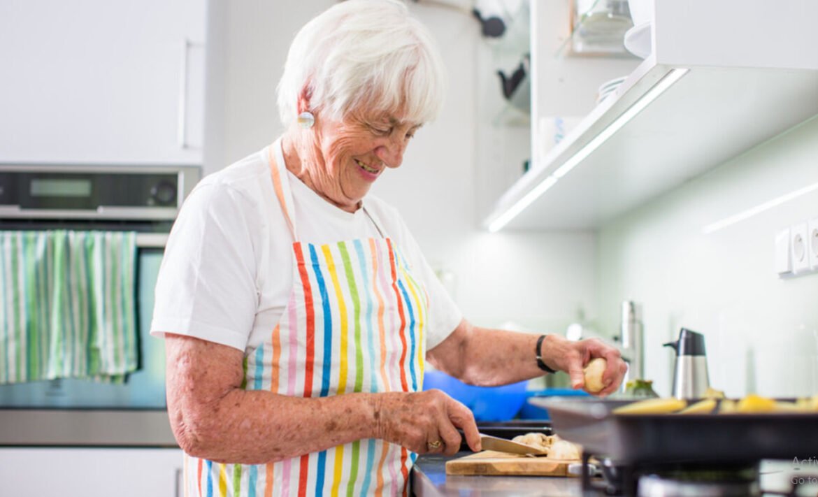 Older woman with white hair chopping on a wooden board in a bright kitchen, peeling a potato.