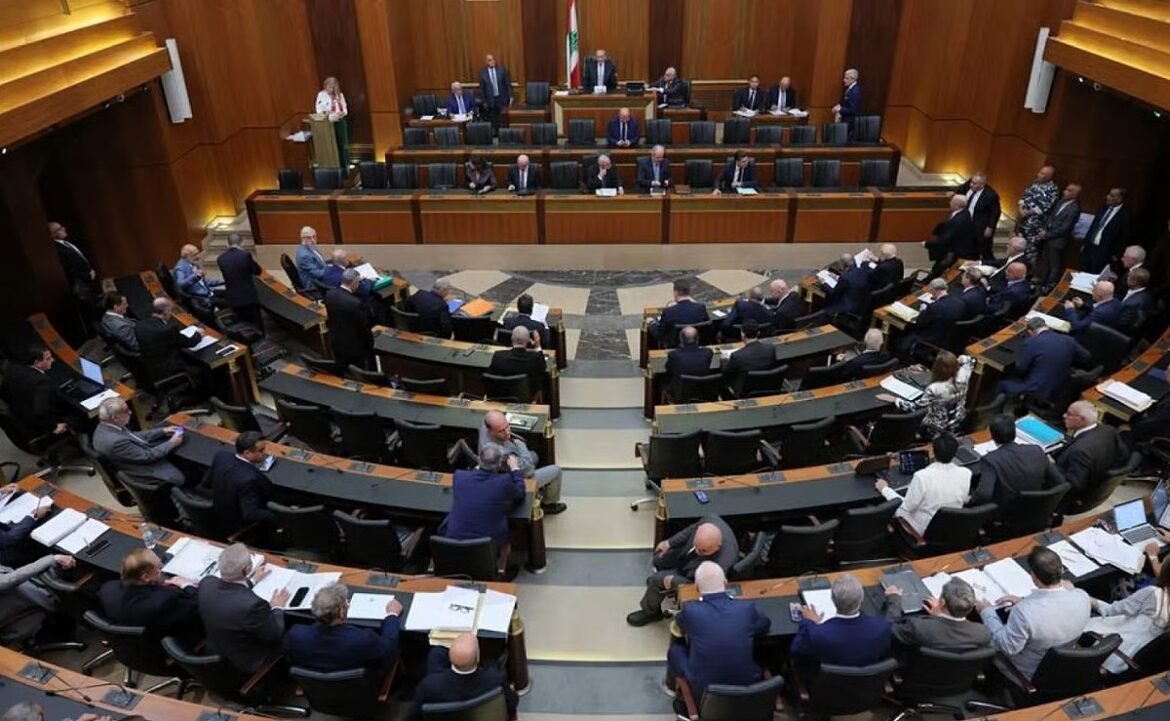 Parliamentary chamber with delegates seated in curved rows, a speaker at the podium, and a flag behind the front desk.