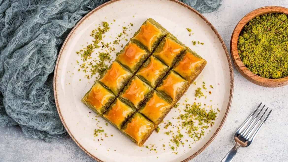 Square rows of pistachio baklava on a white plate, sprinkled with chopped pistachios and a side bowl of pistachio crumble.