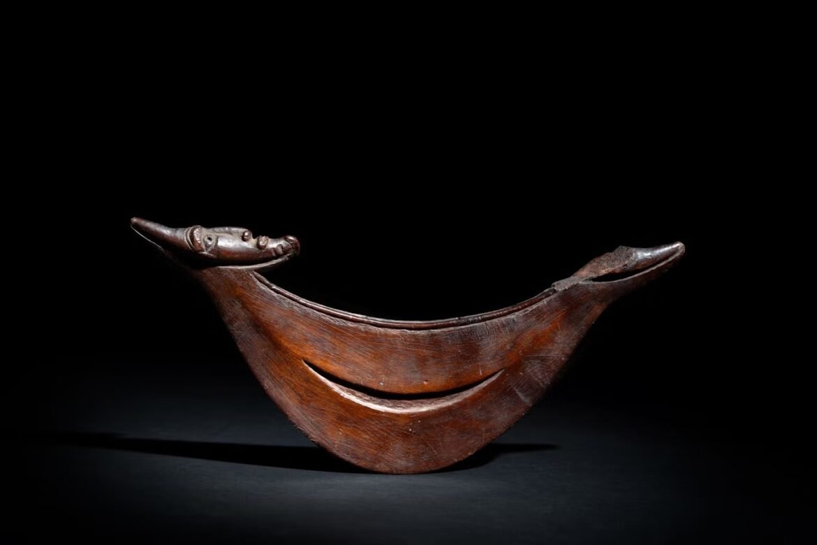 Carved wooden boat-like sculpture with a bird head at the prow against a dark background simplicity in lighting.