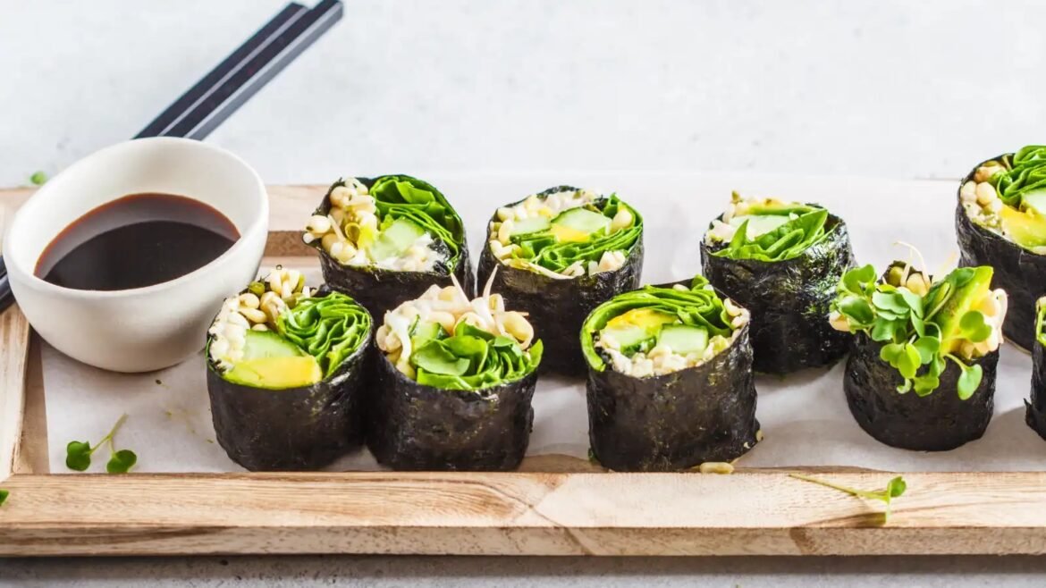 Sushi rolls wrapped in nori on a wooden tray, filled with cucumber and greens, served with a small bowl of soy sauce on the side.