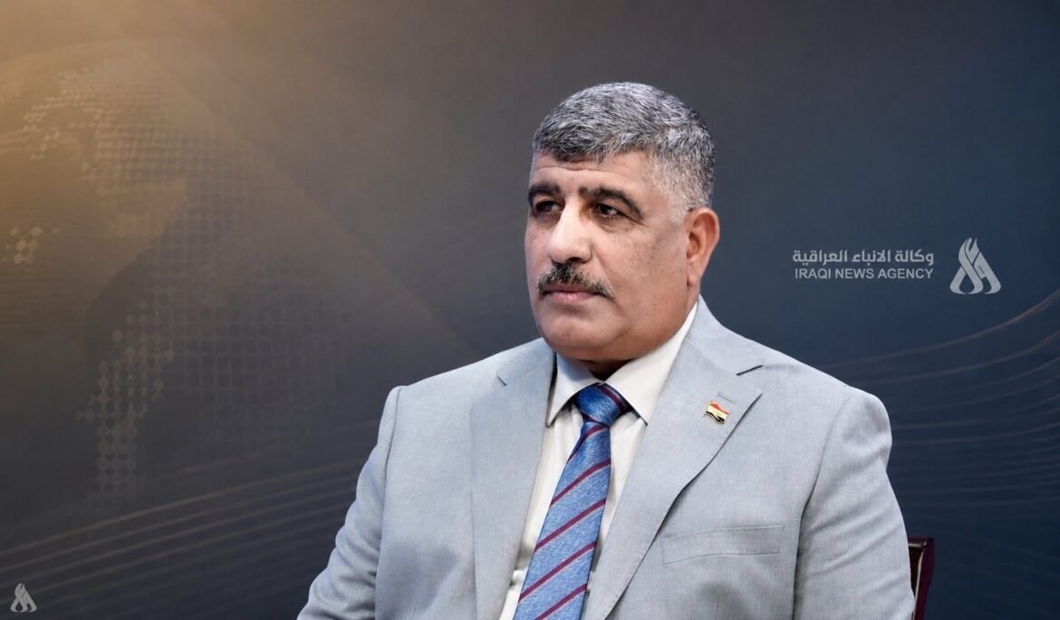 Middle-aged man with gray hair and mustache in a light gray suit and striped blue tie, seated against a dark backdrop with the Iraqi News Agency logo visible on the right.