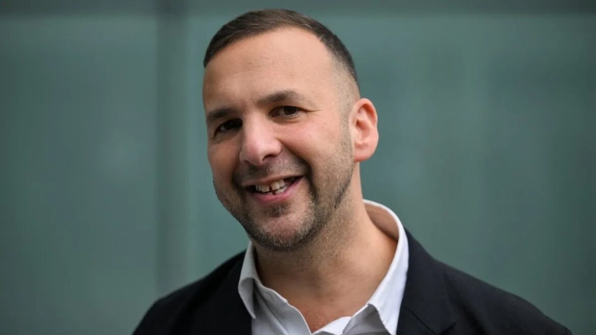 Smiling man in a white shirt and black blazer, facing the camera with a slight head tilt, neutral green background.