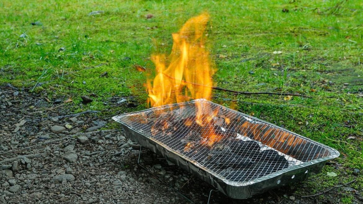 Portable metal grill with burning charcoal on a grassy, Pebble-strewn ground outdoors.