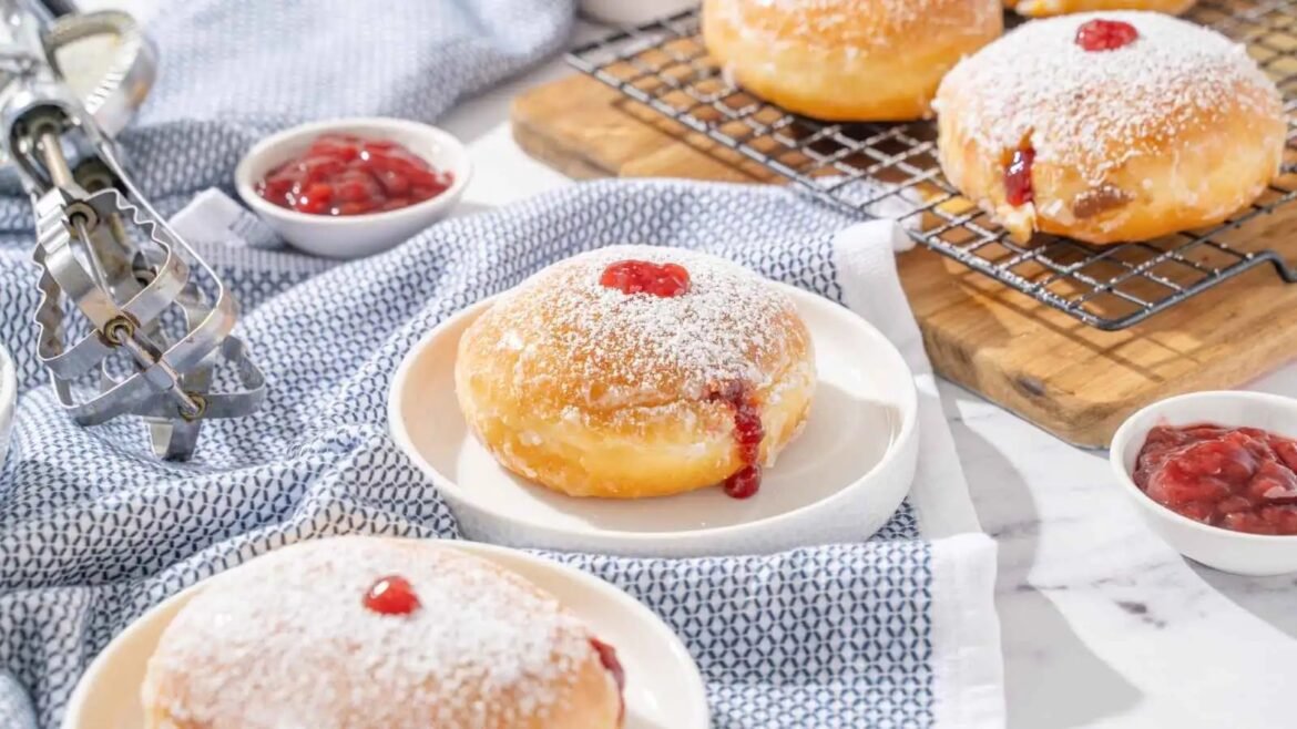 Powdered-sugar doughnuts with red jam centers on plates, with a wire cooling rack and bowls of jam nearby on a striped cloth backdrop.