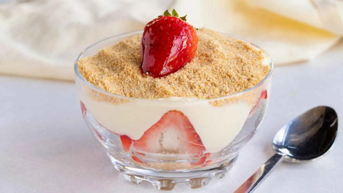 Glass dessert cup layered with vanilla cream, strawberry slices, and a brown crumb topping, topped with a whole strawberry.