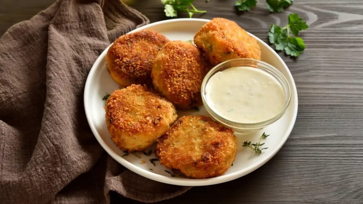Five golden-brown breaded potato croquettes on a white plate with a small bowl of creamy dipping sauce, served on a rustic wooden table with parsley garnish.