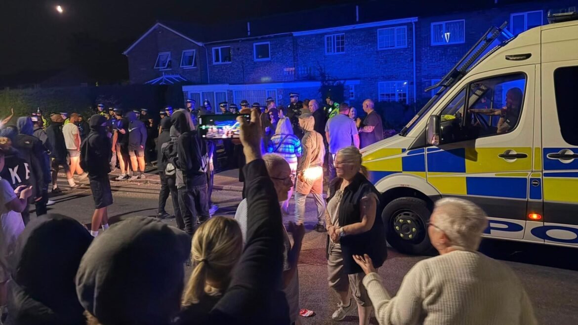 Nighttime street scene with a crowd gathered outside a residential building and an illuminated police/emergency vehicle nearby. People watch and take photos as blue lights flash.