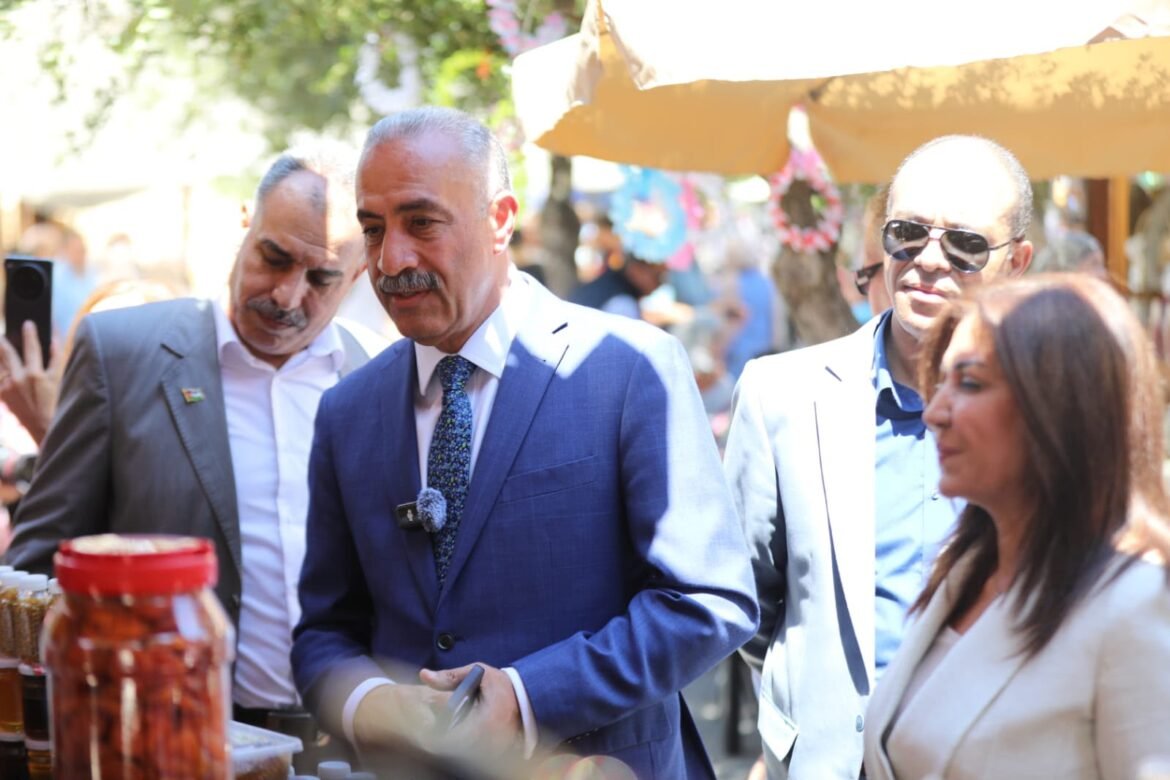 Man in a blue suit and patterned tie stands at an outdoor market stall, with jars of preserves visible in front and other people nearby.