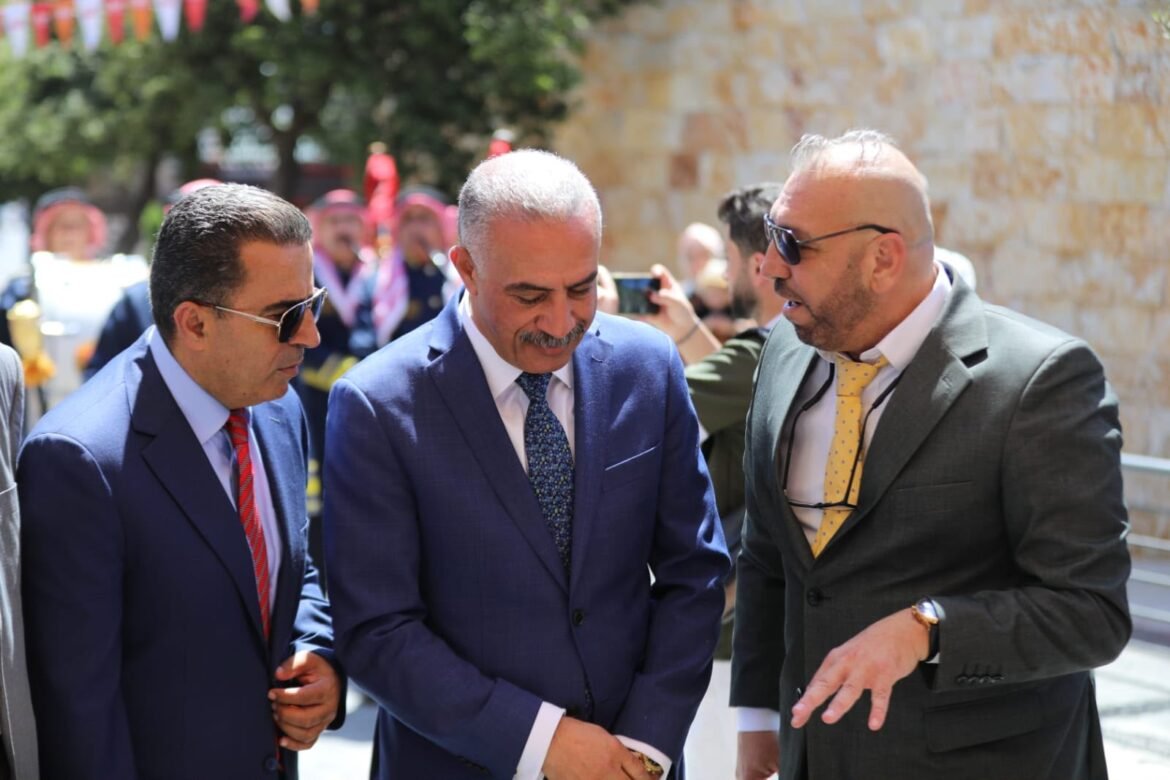 Three suited men in conversation outdoors, middle man looking down as the others speak, background stone wall and onlookers.