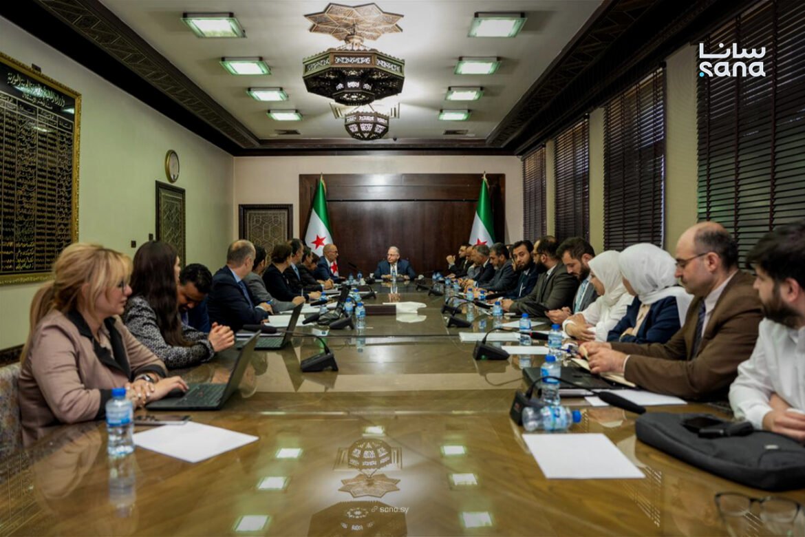 Diplomatic meeting around a long conference table with laptops and water bottles; flags in the background.