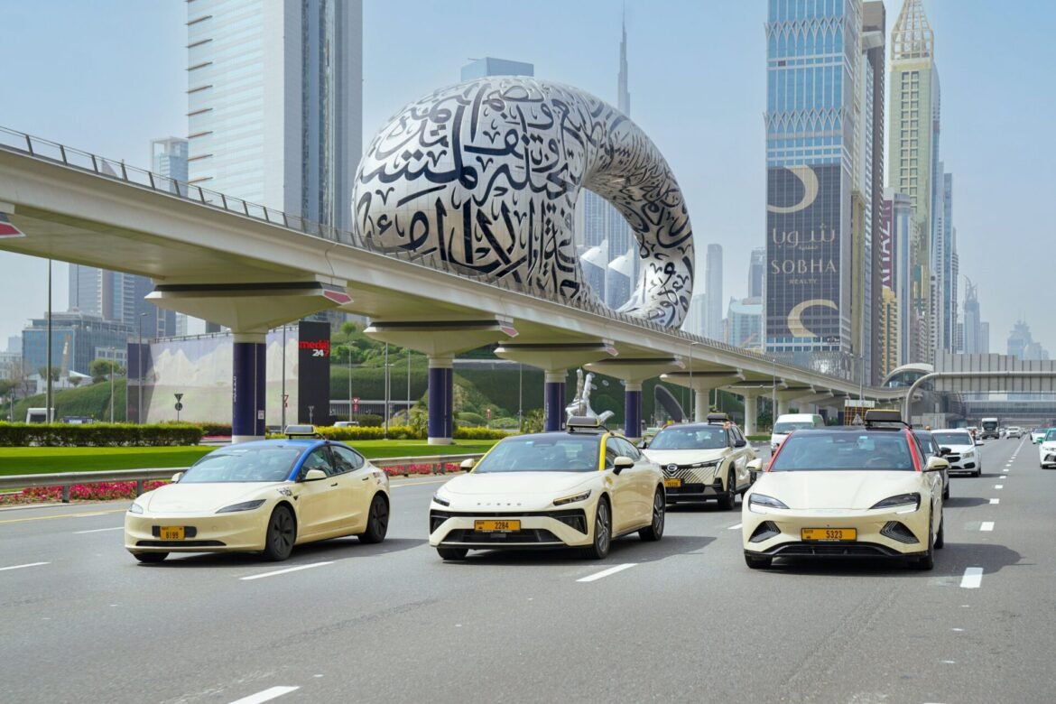 Cars drive on a multi-lane road beneath an elevated track, with a giant white loop sculpture covered in Arabic calligraphy and tall skyscrapers behind.
