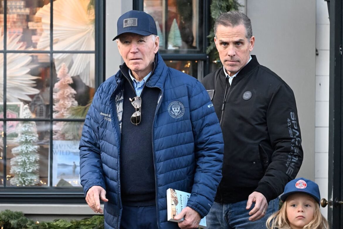 Two adult men and a young girl stand outside a shop with holiday window displays; one man in a blue puffer jacket and cap, the other in a black jacket, girl in a blue cap nearby.
