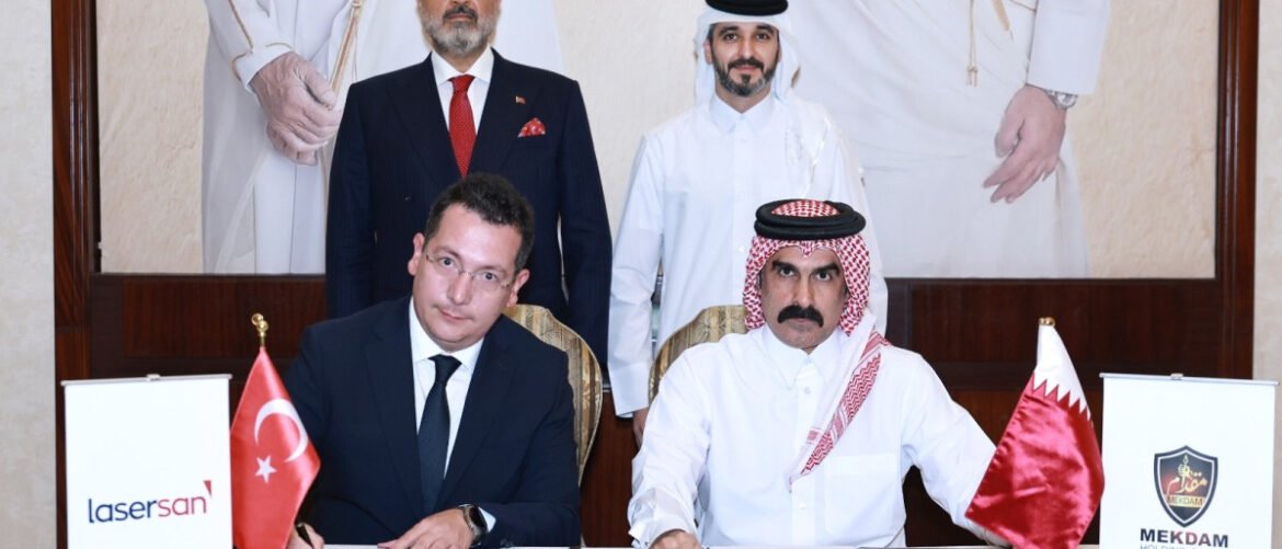 Two men sign an agreement at a formal ceremony, with Turkish and Qatari flags beside them and suited officials behind.