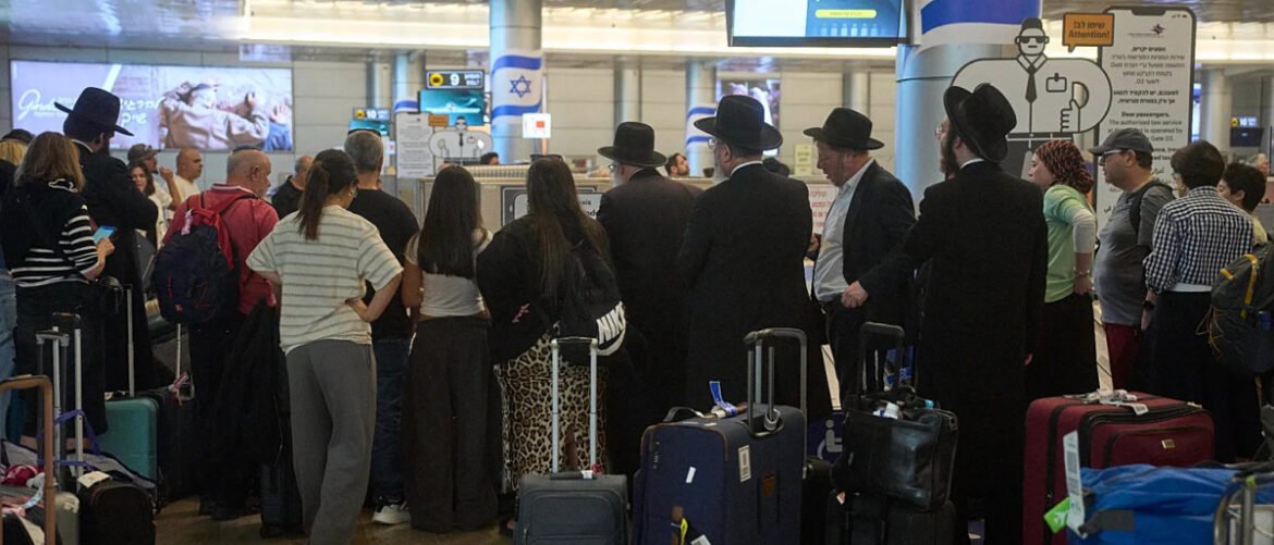 Crowd of passengers waiting in line with rolling luggage at an airport security checkpoint; some men wear traditional black hats, Israeli flag visible in the background.