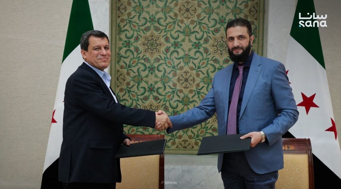 Two men in suits shake hands in front of ornate backdrop, each holding a folder as they seal an agreement.