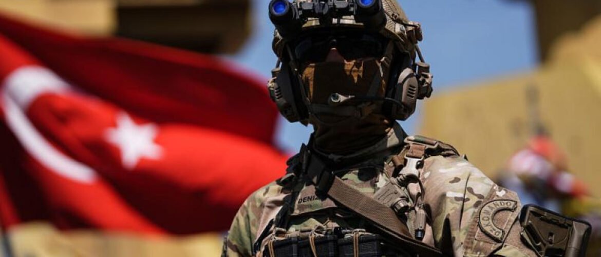 Soldier in full tactical gear with night-vision goggles, stance in front of a Turkish flag.